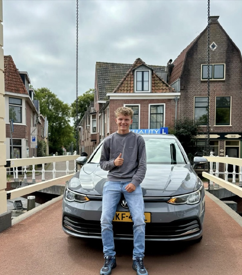 Binc met duim omhoog voor de grijze VW Golf-lesauto van Quality op een brug in Alkmaar
