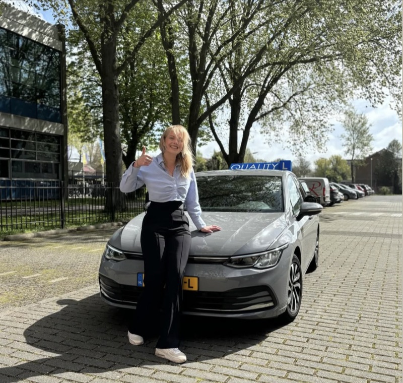 Yinka met duim omhoog voor de grijze VW Golf-lesauto van Quality
